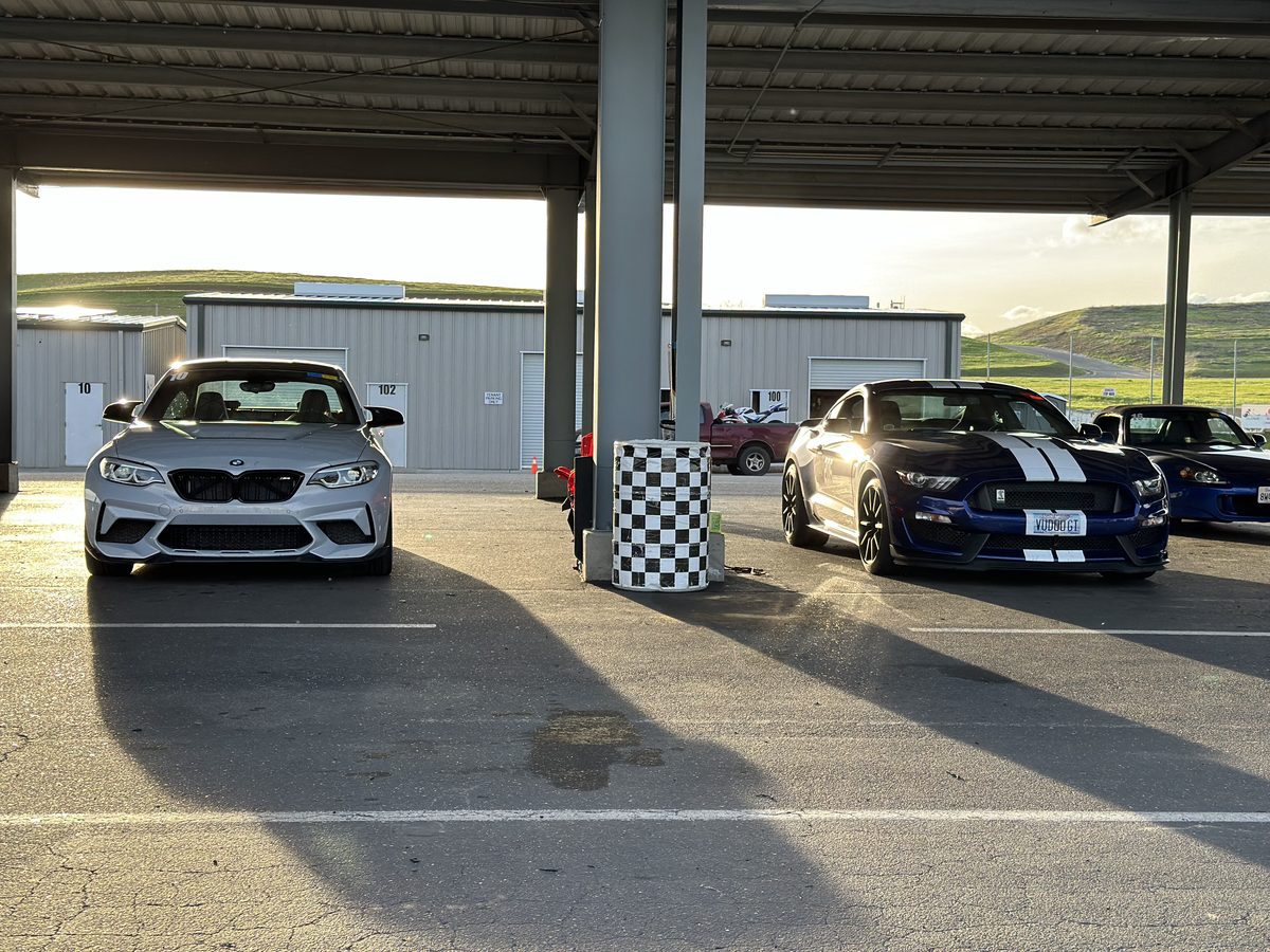 Silver BMW M2 and a blue Shelby GT350 with white racing stripes parked in the track paddock